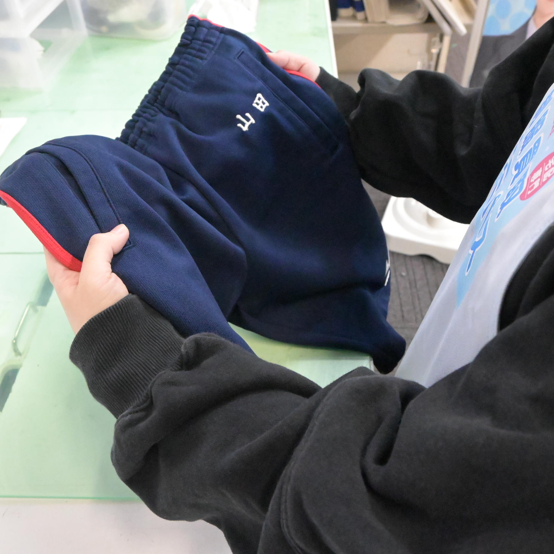 ラミパススタッフがジャージを両手で持ち、「山田」のネーム刺繍箇所を確認している