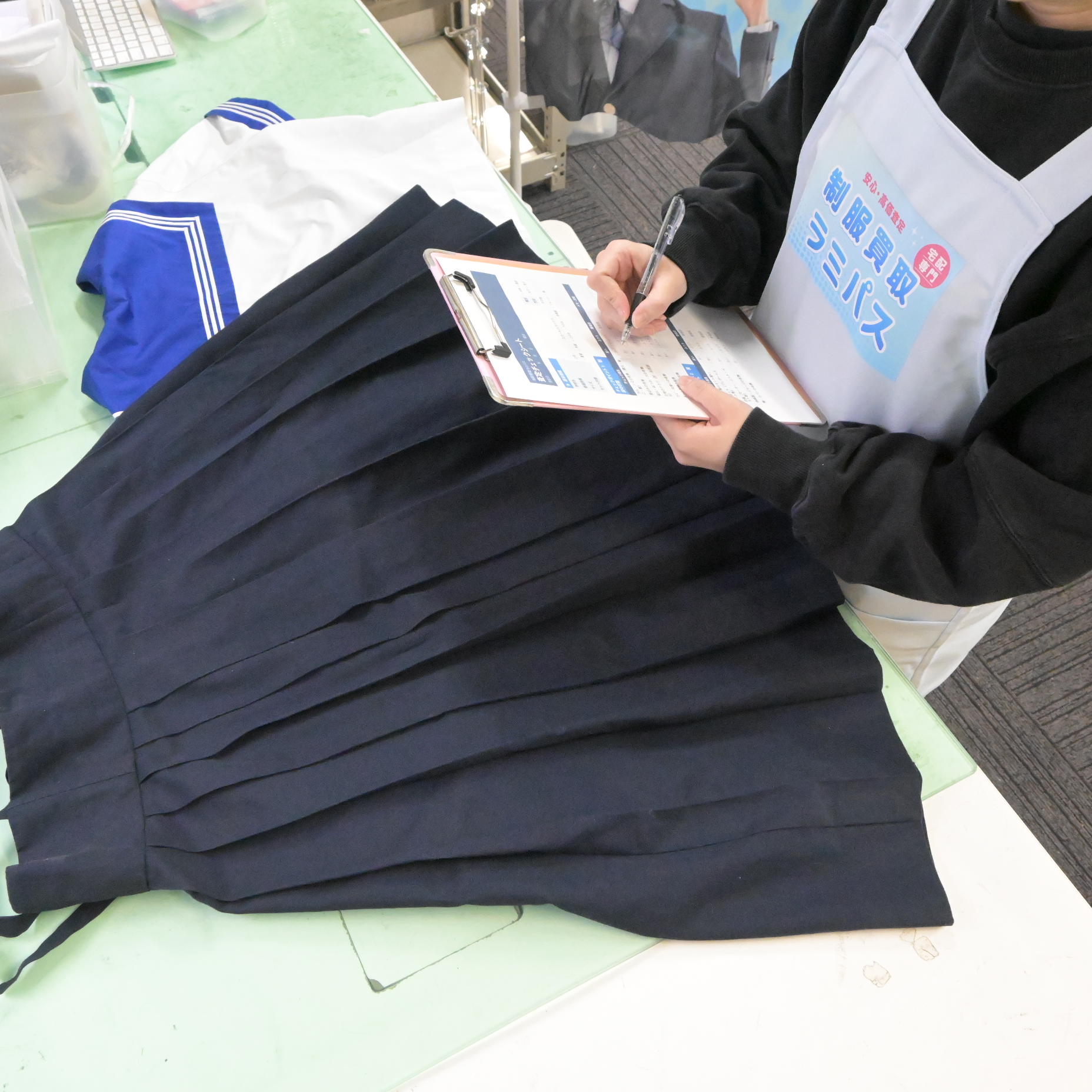 制服買取ラミパスのスタッフがクリップボードで制服スカートを丁寧に査定している様子。ブランドエプロン着用の専門スタッフが一点ずつ状態を確認する