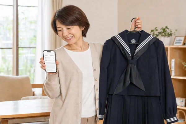 学習院女子中等科の制服をハンガーで持ちスマートフォンで買取査定を確認する母親
