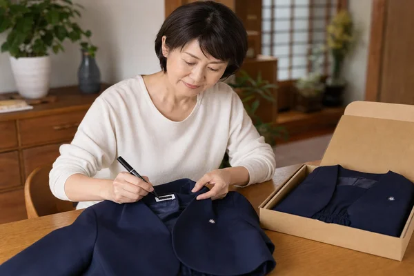 聖霊高校の制服の衿ネームタグを油性ペンで塗りつぶし、ダンボールに梱包して宅配買取の準備をする母親