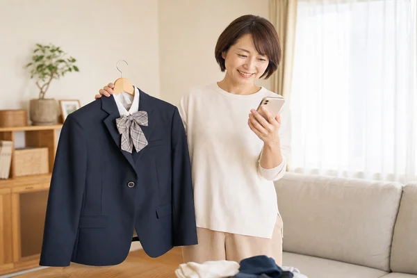 東京家政大学附属女子の制服をハンガーで持ちスマートフォンで買取査定を確認する母親