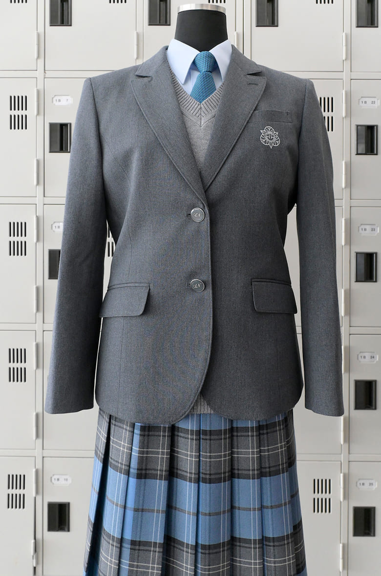秋田県立秋田西高等学校の女子新制服(令和8年度導入)。グレーのブレザーにブルー系チェックのプリーツスカート、グレーのVネックセーター、ブルーネクタイを合わせたブレザースタイル