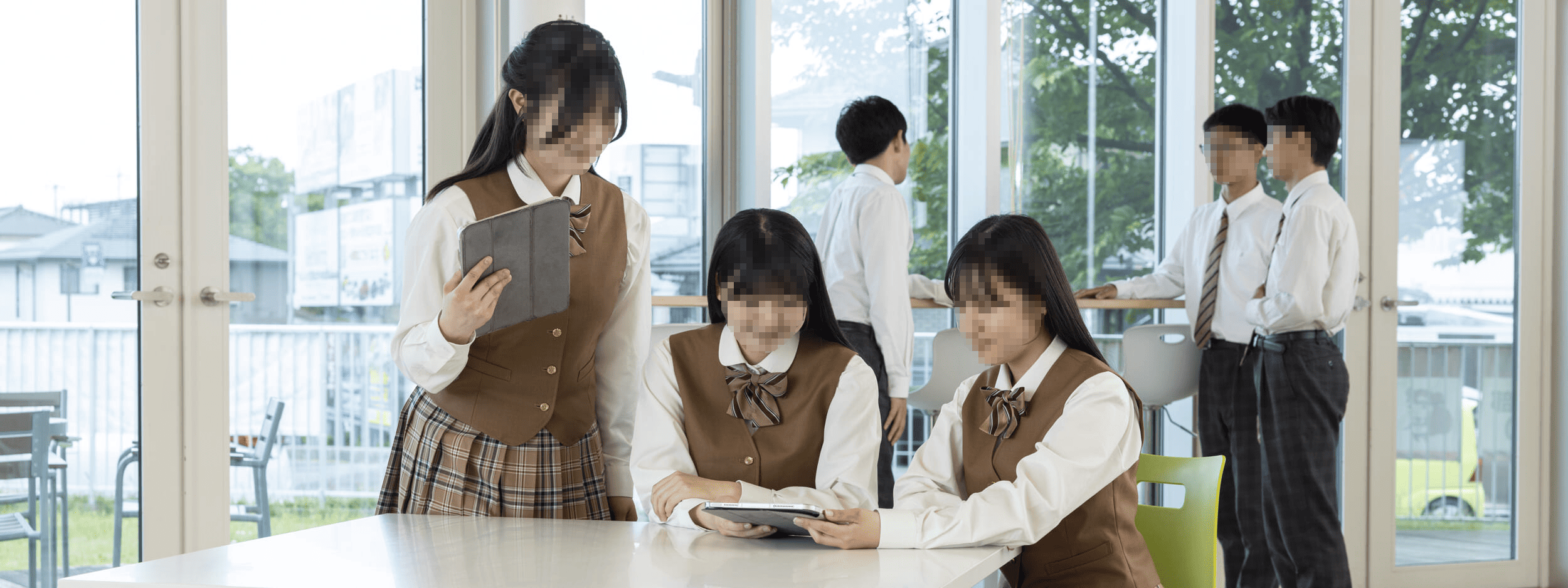 作新学院高等学校の制服姿の女子生徒がタブレットで学習しているシーン。ブラウンベスト・チェックスカートの女子制服と白シャツ・ネクタイの男子制服。