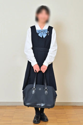 京都聖母学院中学校の冬服。濃紺のジャンパースカートに白の長袖ブラウス、水色のリボン、指定バッグを持った女子生徒